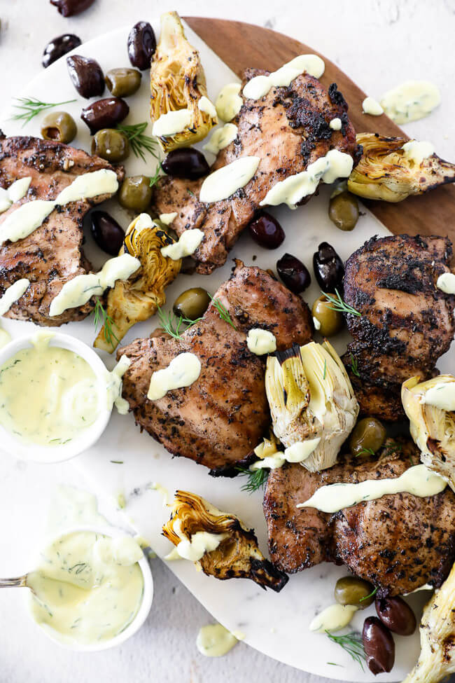 Overhead image of grilled mediterranean chicken on a platter with sauce drizzle, artichoke hearts and olives.