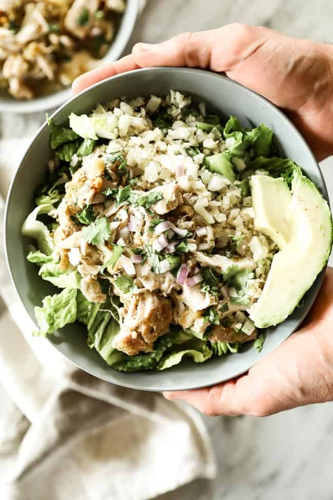 Holding a bowl of Mexican shredded chicken with romaine lettuce, caulirice, shredded chicken, avocado and fresh chopped red onion and cilantro. 