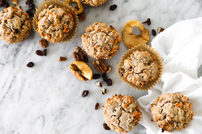 Horizontal overhead image of morning glory breakfast muffins spread out on marble with dried apples, raisins and pecans sprinkled around. 