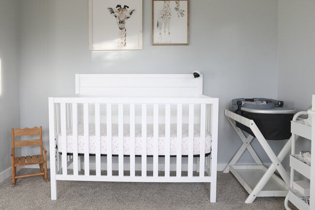 Horizontal image of white baby crib in nursery. Giraffe wall art above crib and bassinet and mini rocking chair off to the sides. 