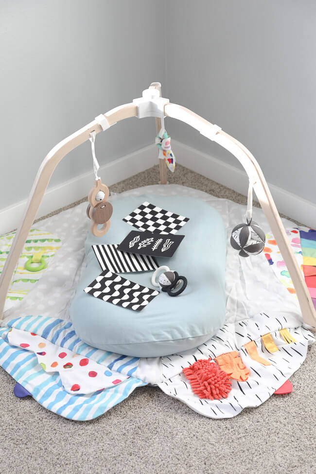 Vertical image of baby play gym on the floor with an infant lounger and extra toys laying on top. 