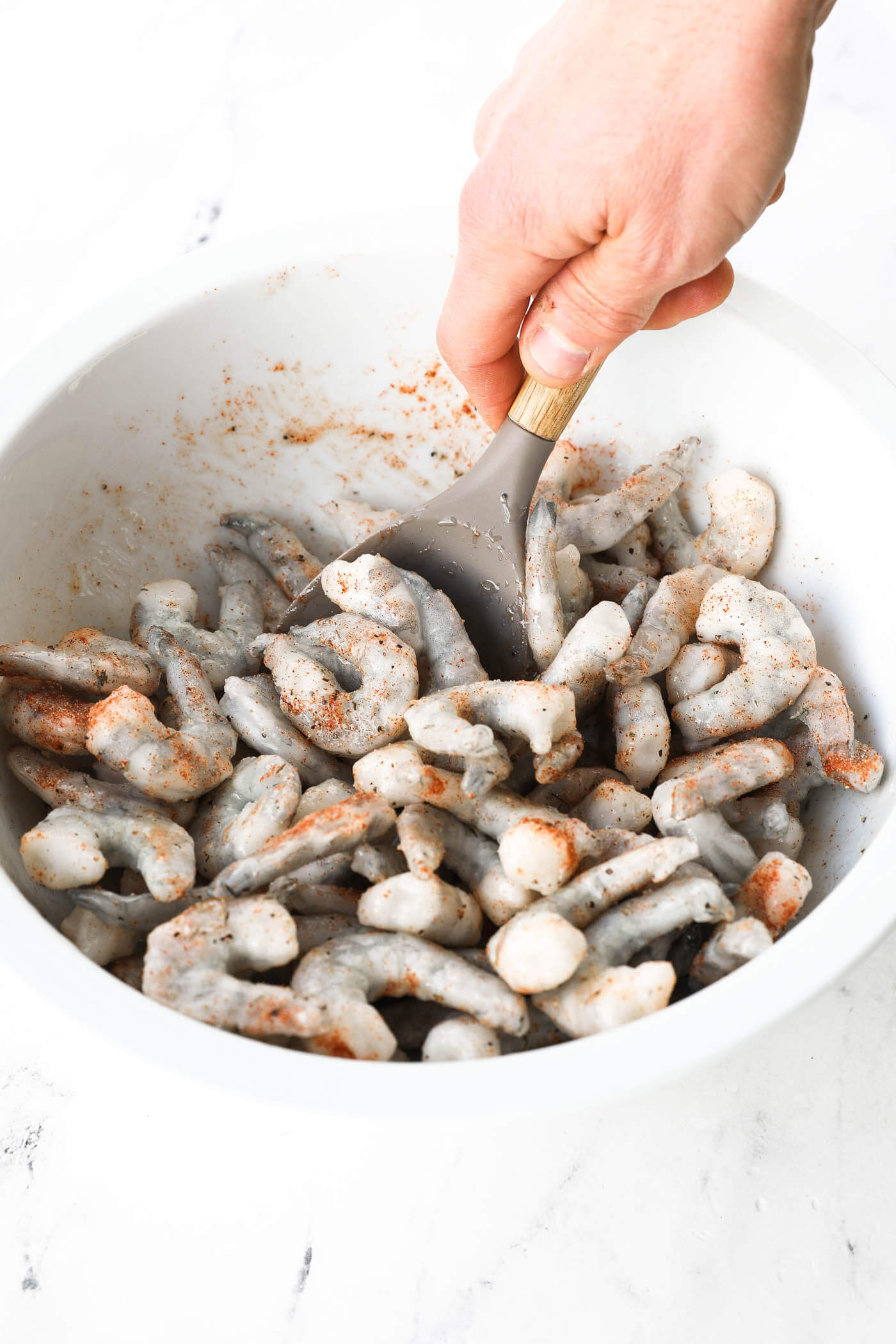 A hand mixing the seasonings with the oiled shrimp in a bowl.
