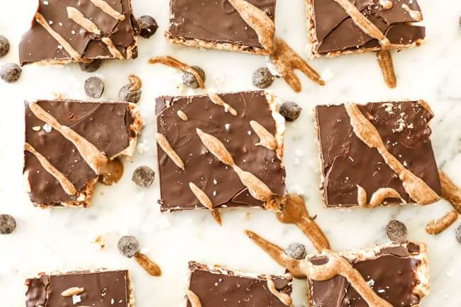 Peanut butter bars lined up on marble with extra chocolate chips and sea salt spread all around. Drizzled with nut butter on top. 