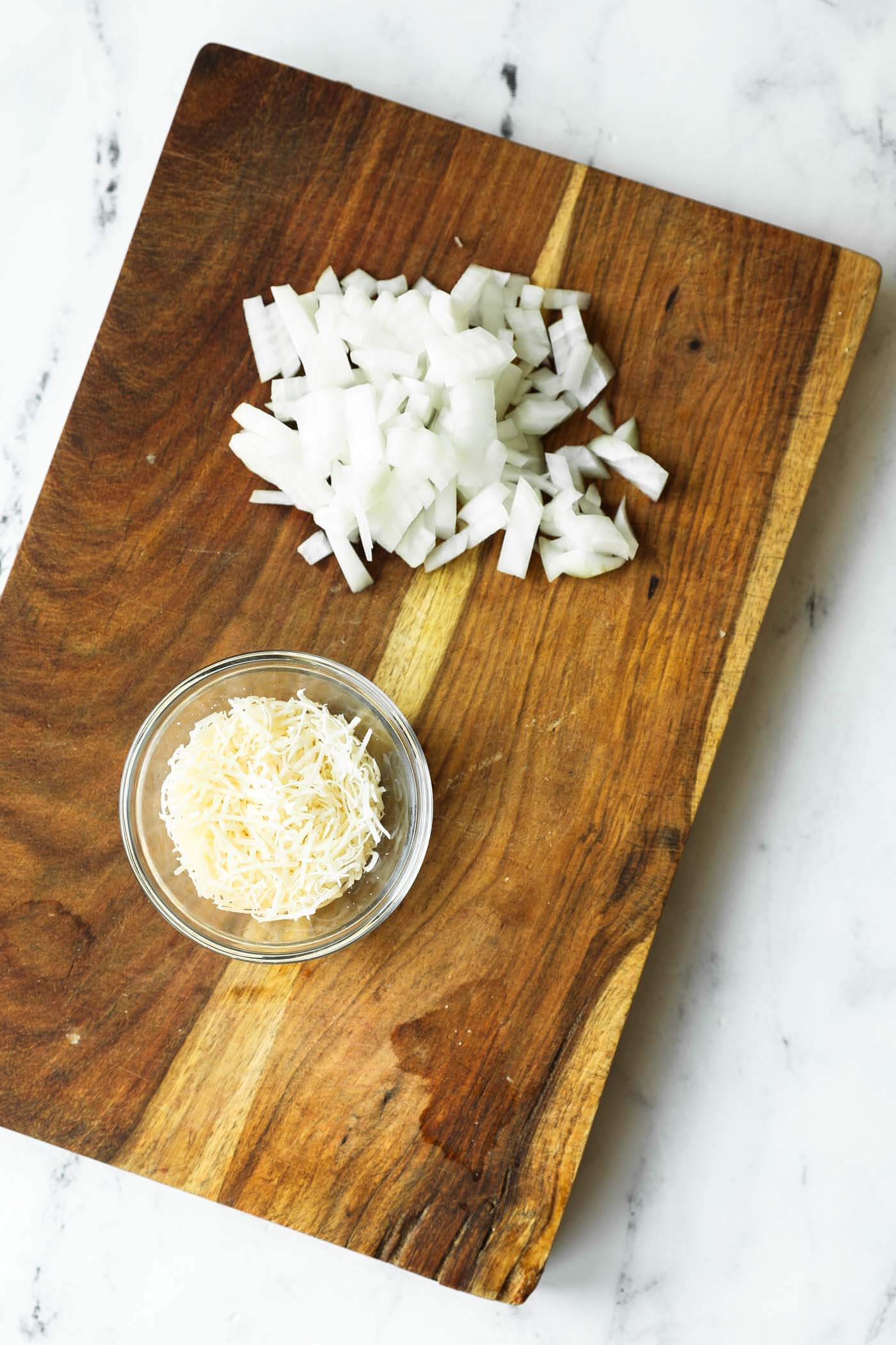 Chopped white onions and a small glass bowl of shredded cheese sit on a wooden cutting board on a marble countertop.
