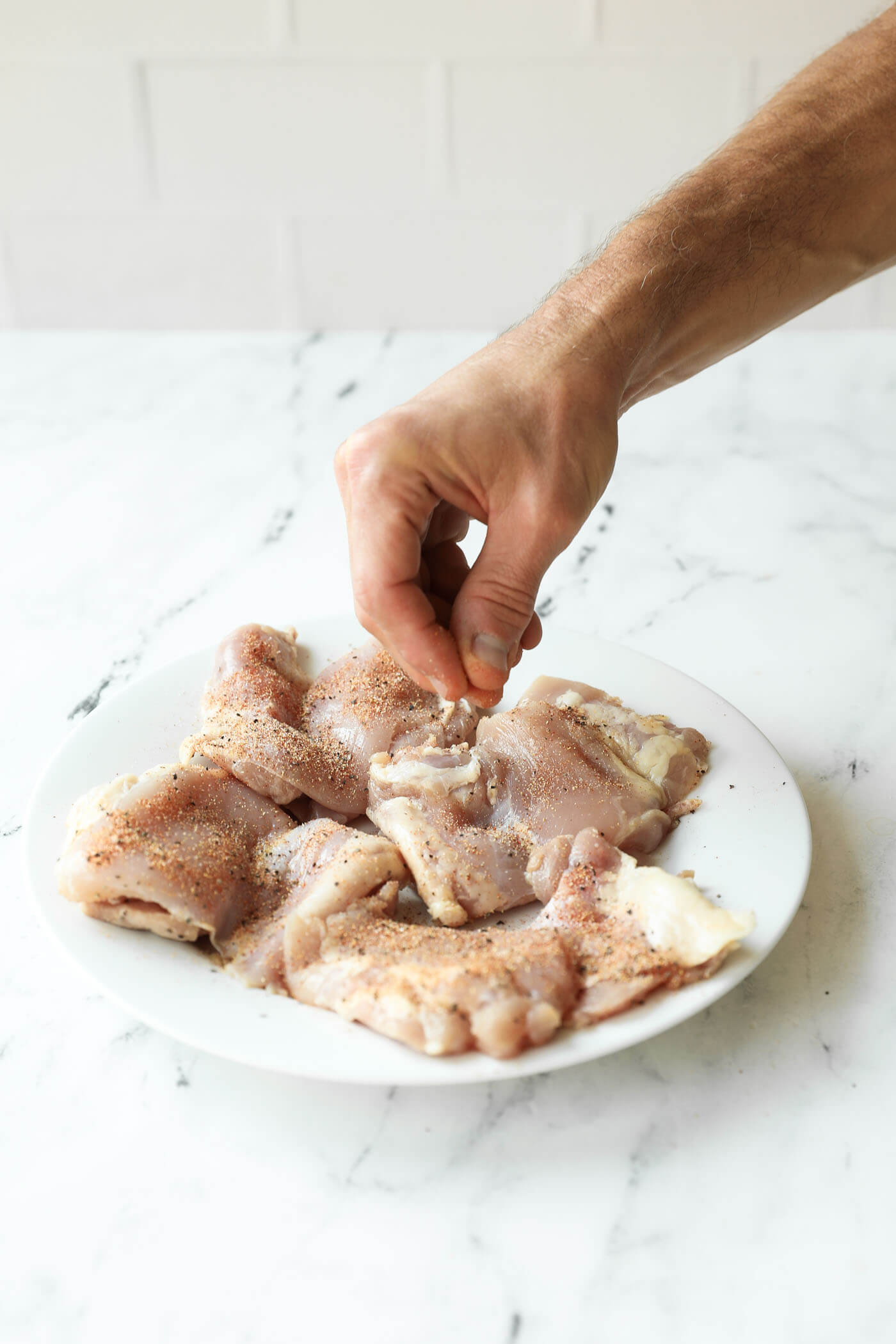 A hand sprinkles seasoning onto raw chicken pieces arranged on a white plate on a marble countertop.