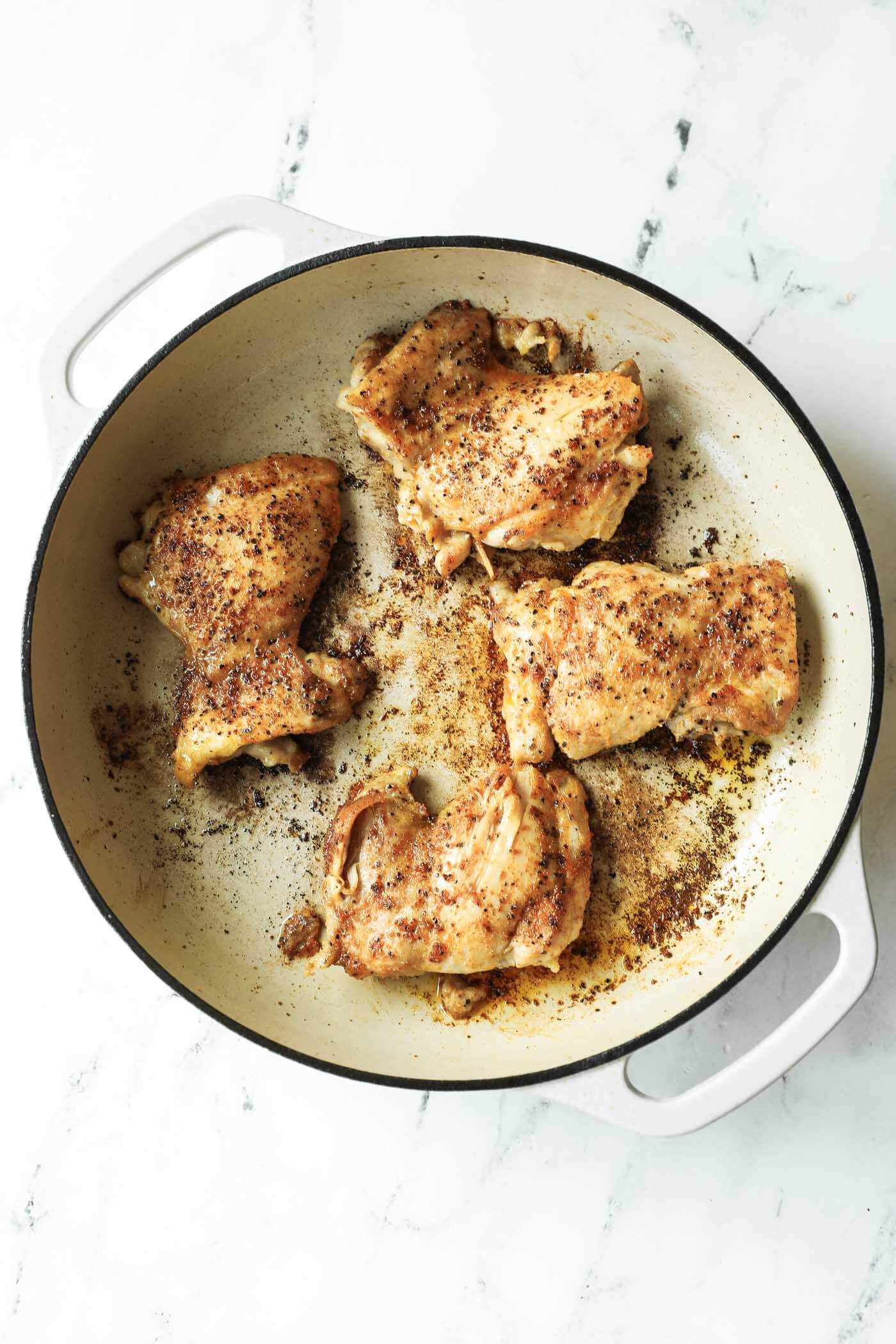 Four seasoned chicken thighs are browning in a white enameled pan on a marble surface.