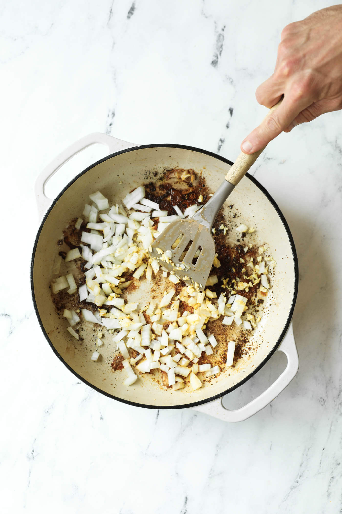 A hand holding a spatula stirs chopped onions and garlic in a white pot on a white marble surface.