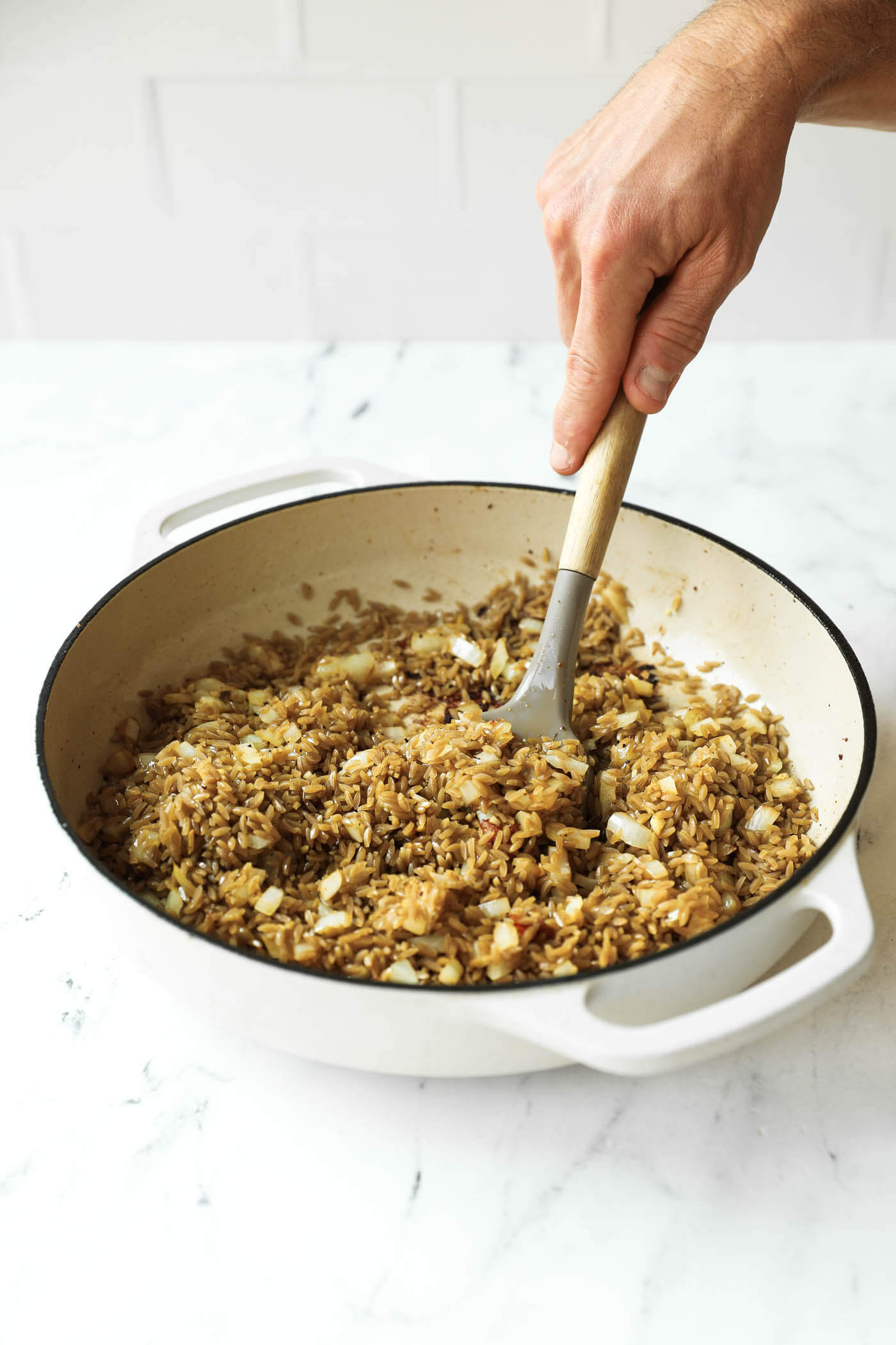 A hand stirs cooked rice and diced onions in a white saucepan on a marble countertop.
