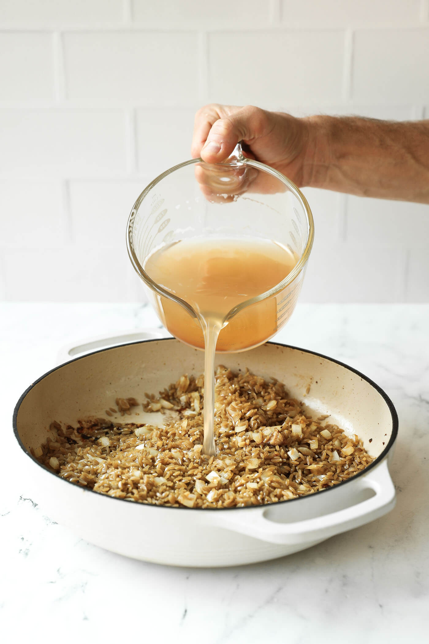 A hand pours broth from a glass measuring cup into a white pan with sautéed onions and grains on a marble countertop.