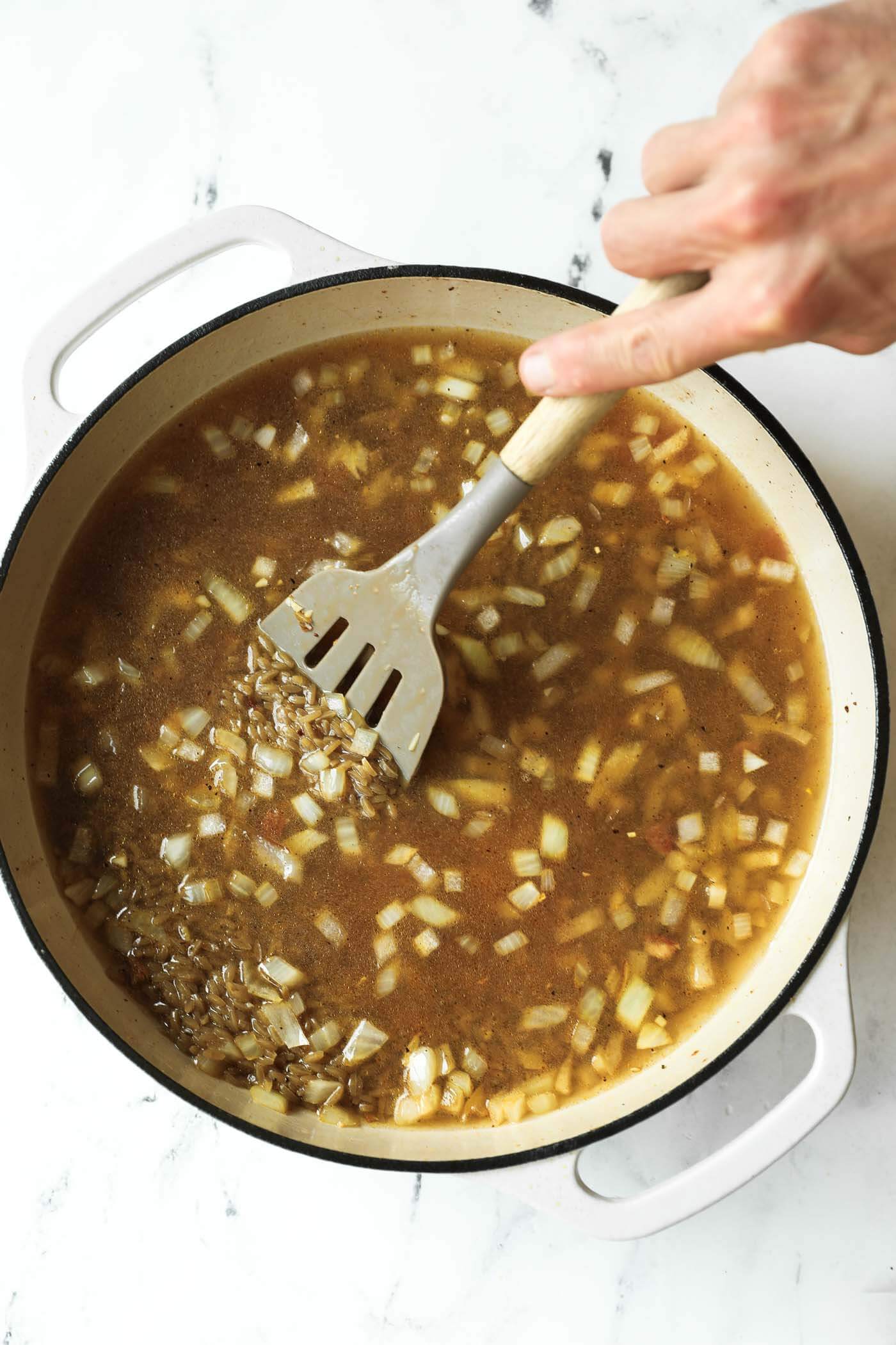 A hand stirs chopped onions and broth with a spatula in a large white pot on a marble surface.