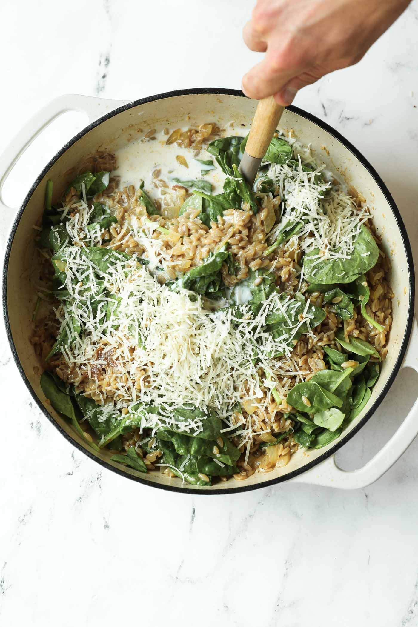 A hand stirring spinach, grated cheese, and creamy sauce into cooked orzo in a white pot on a white surface.
