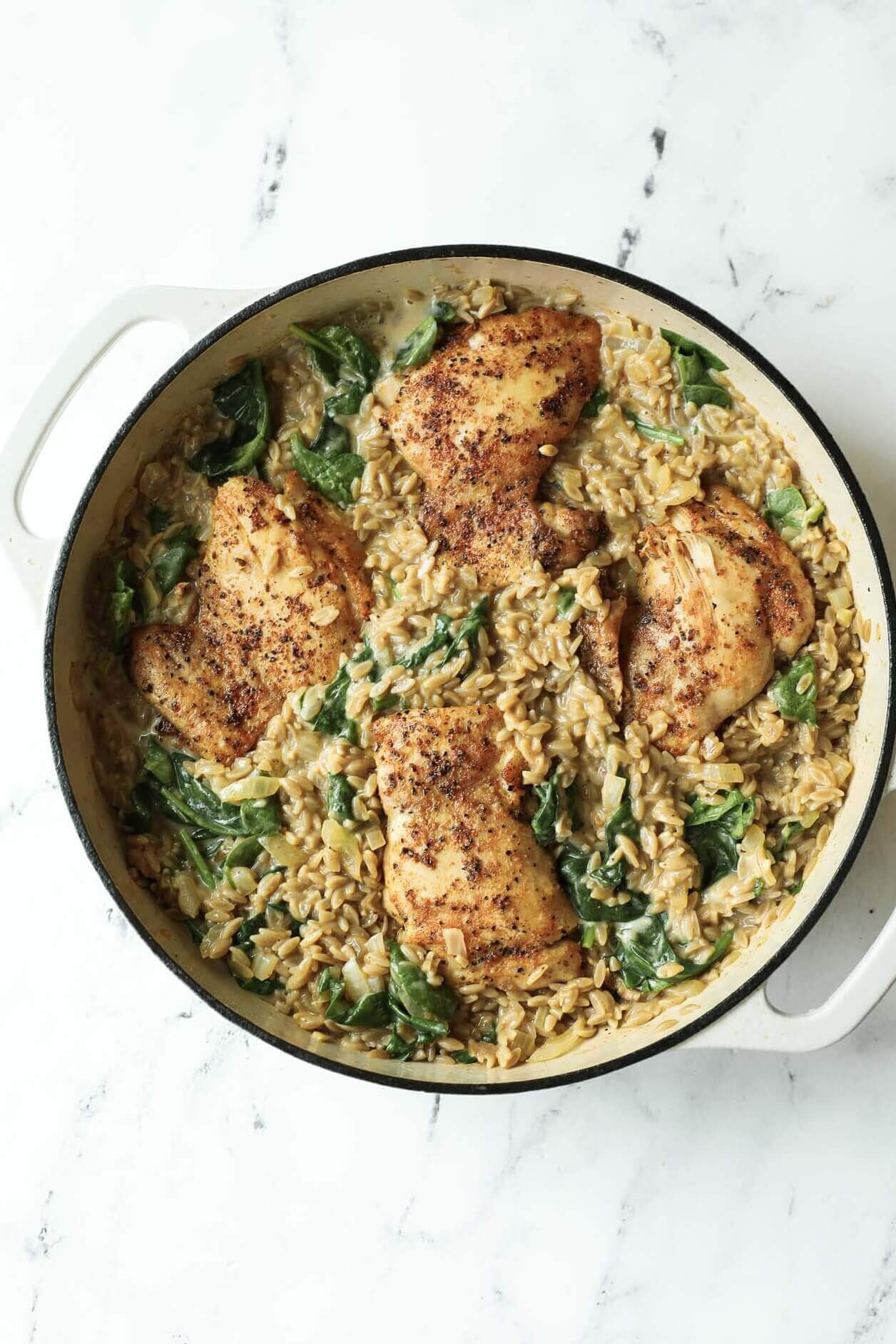 A white pot filled with seasoned chicken thighs, creamy rice, and spinach, placed on a white marble surface.