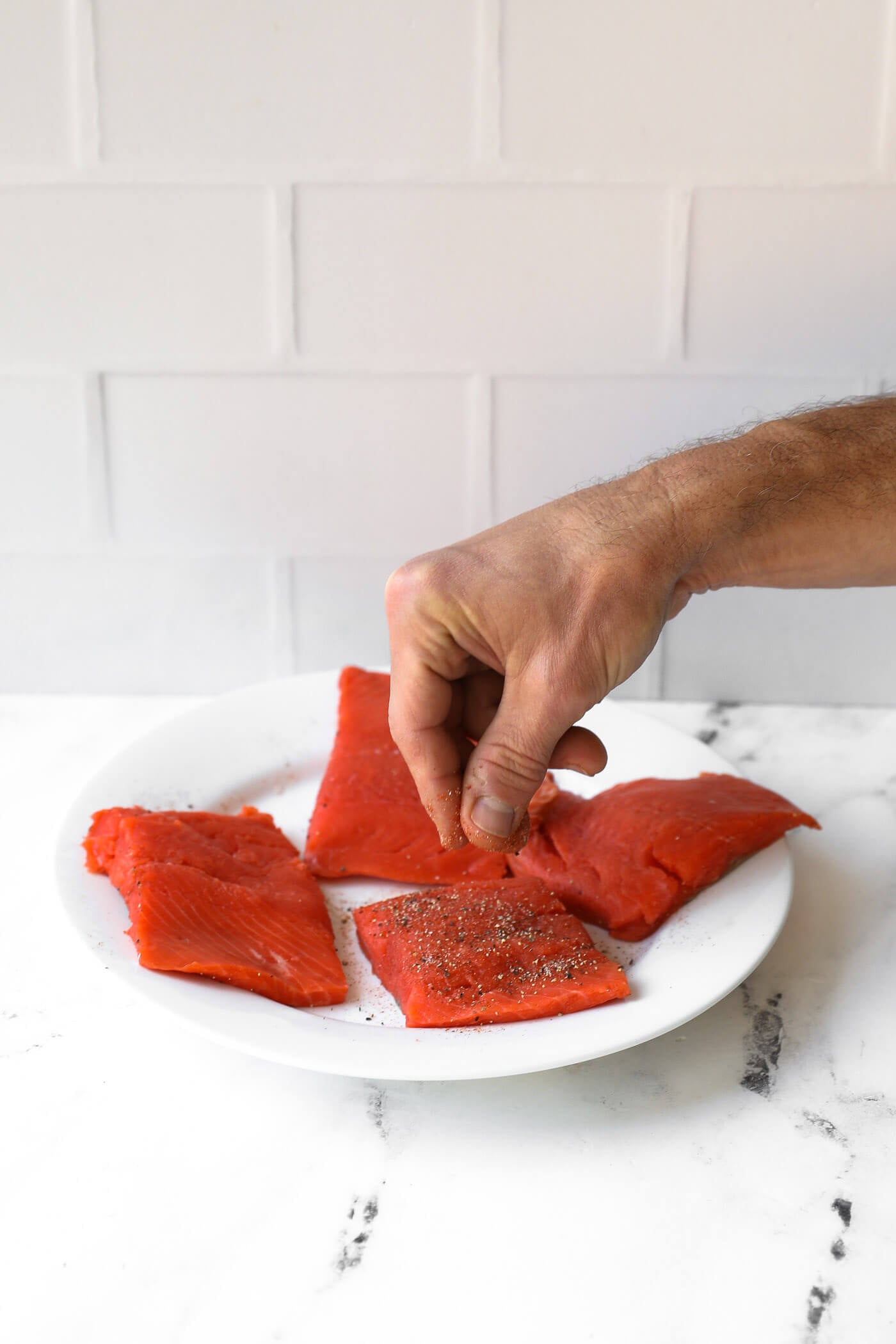 A hand sprinkles seasoning onto four raw salmon fillets arranged on a white plate, set on a marble countertop with a white tiled backsplash.