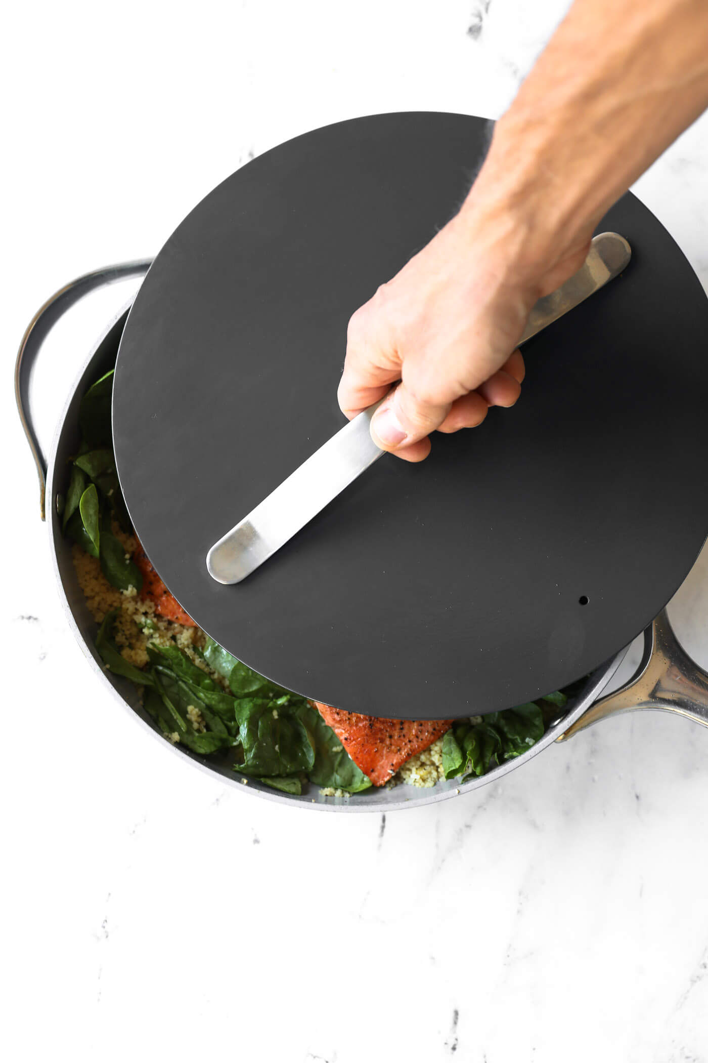 A hand lifts the lid from a pot containing seasoned salmon, spinach, and grains, set on a white surface.