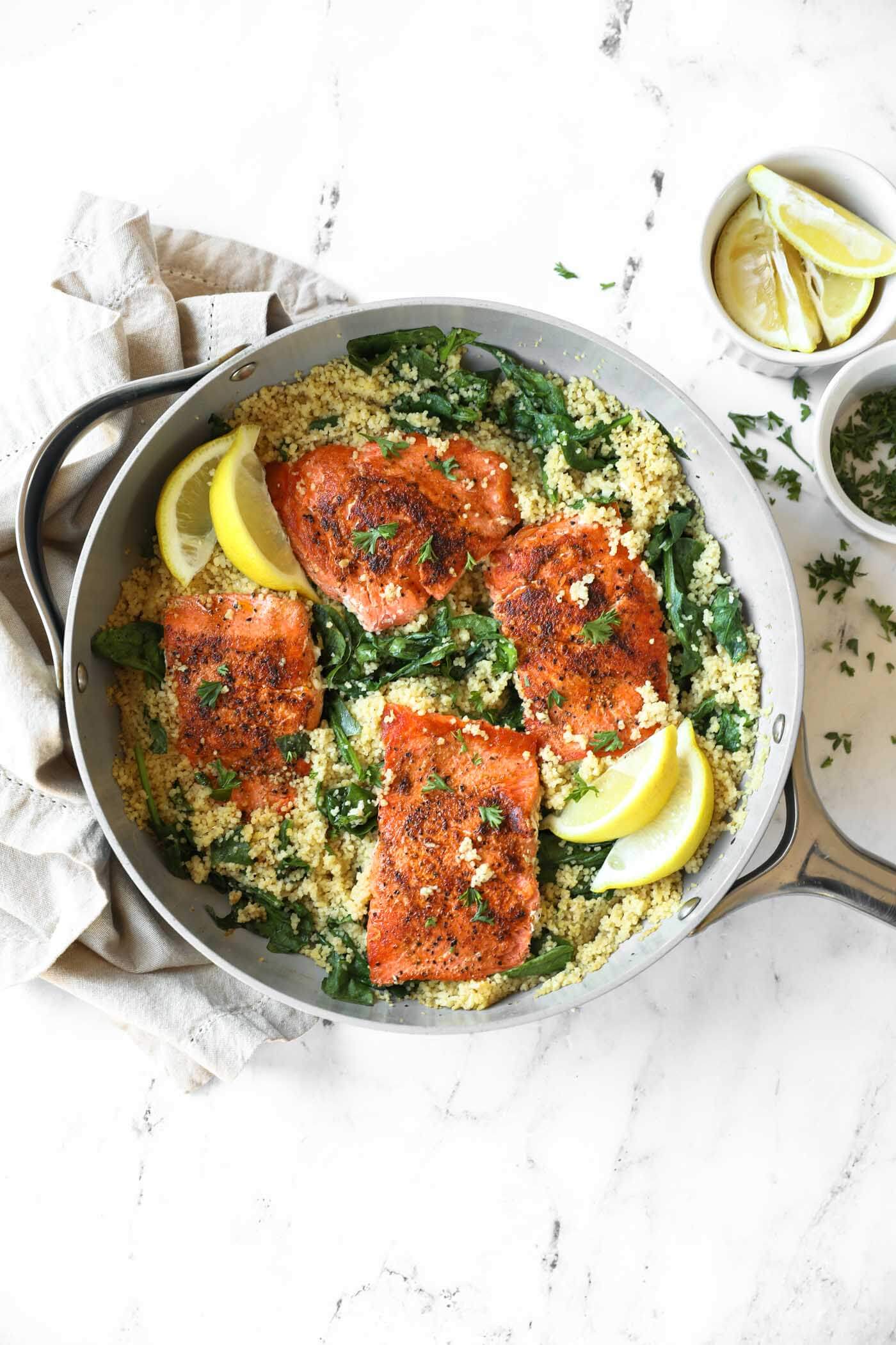 A skillet with four pieces of seasoned salmon on a bed of couscous and greens, garnished with lemon wedges and fresh herbs. A bowl of lemon slices sits nearby.