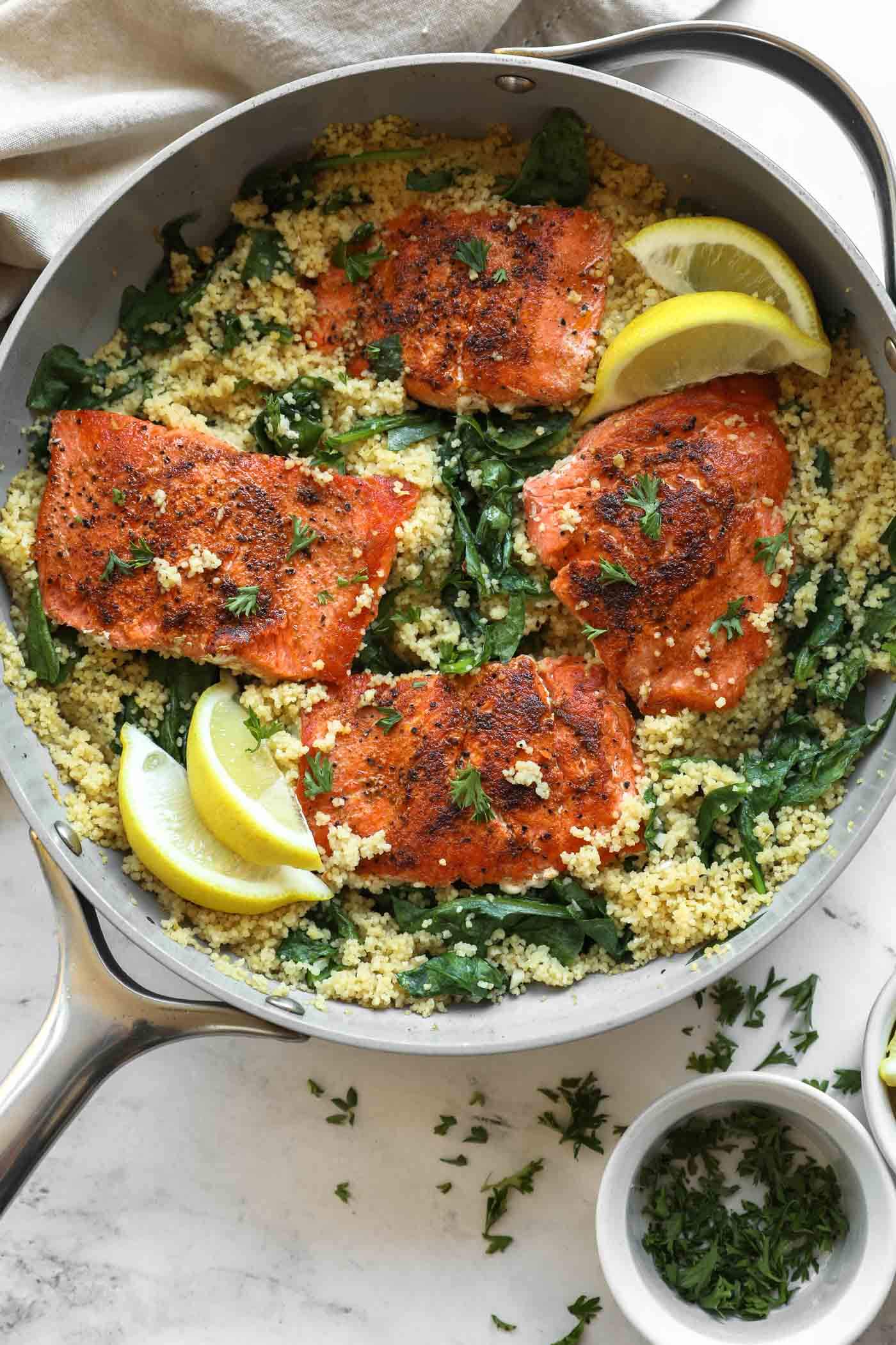 Four pieces of seasoned salmon on a bed of couscous with spinach in a skillet, garnished with lemon wedges and chopped herbs.