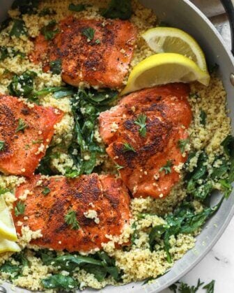 Four seasoned salmon fillets on a bed of couscous and spinach in a skillet, garnished with parsley and lemon wedges.