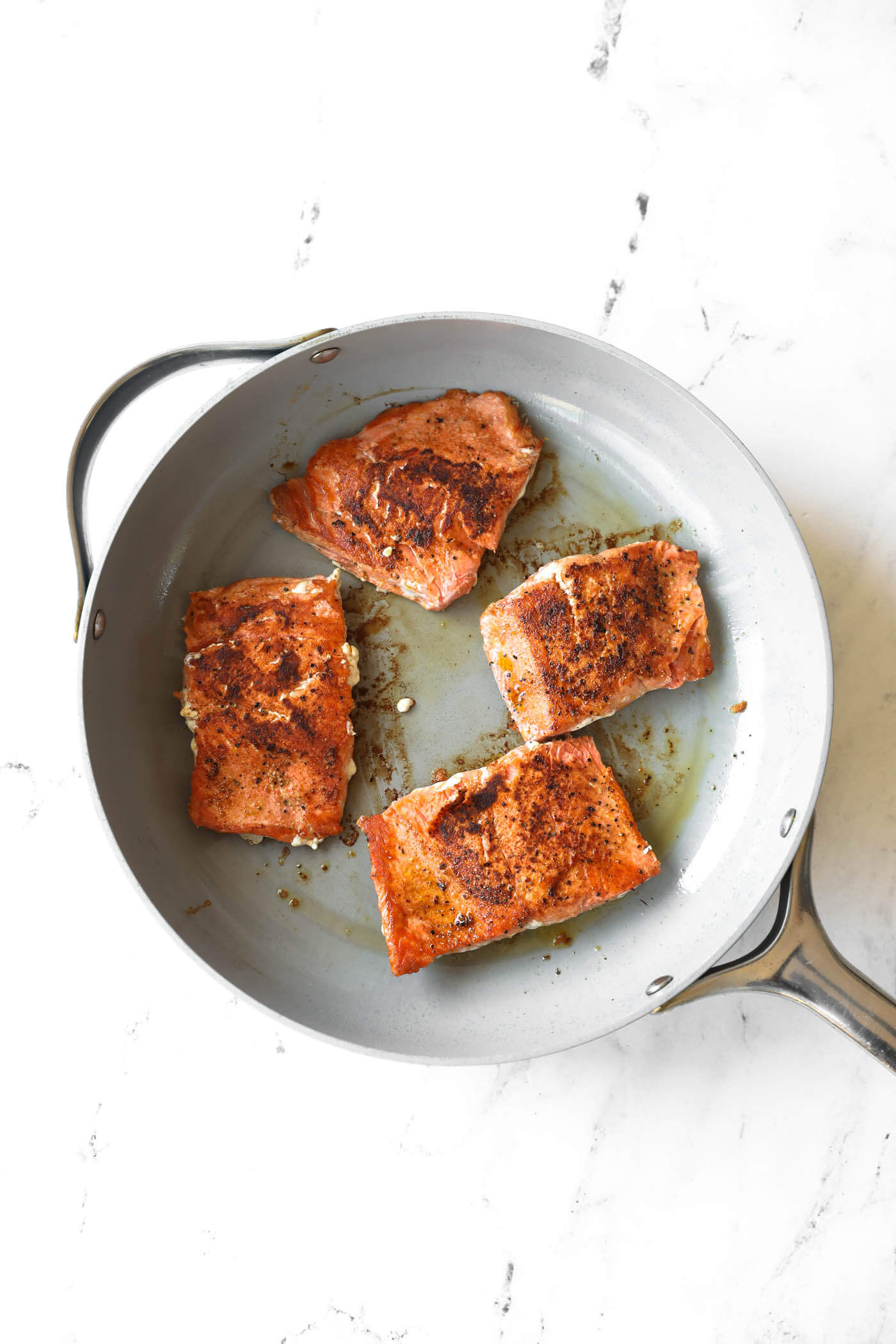 Four seasoned salmon fillets are cooking in a gray skillet on a white surface.