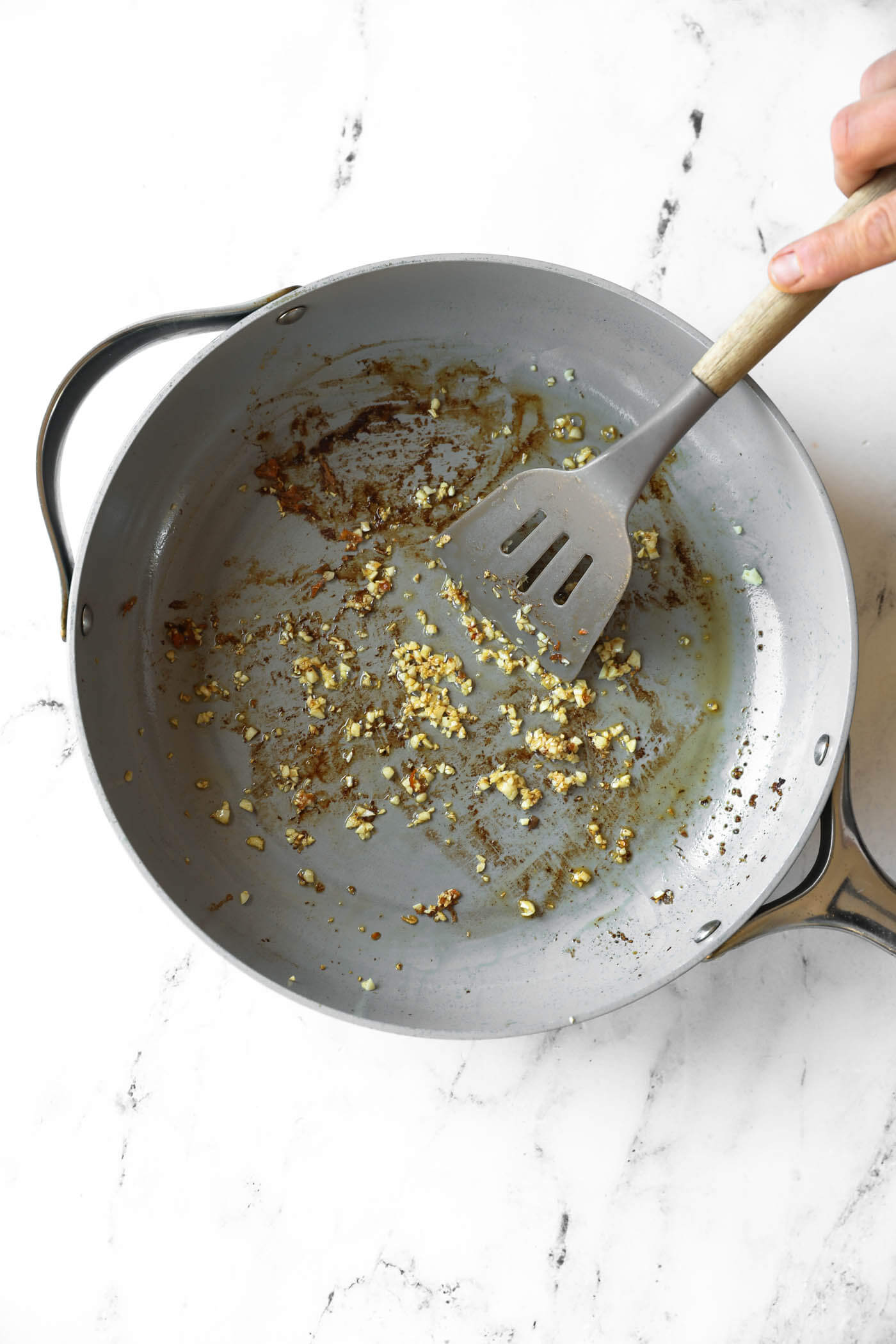 A hand holding a spatula stirs minced garlic browning in a large skillet on a white marble surface.