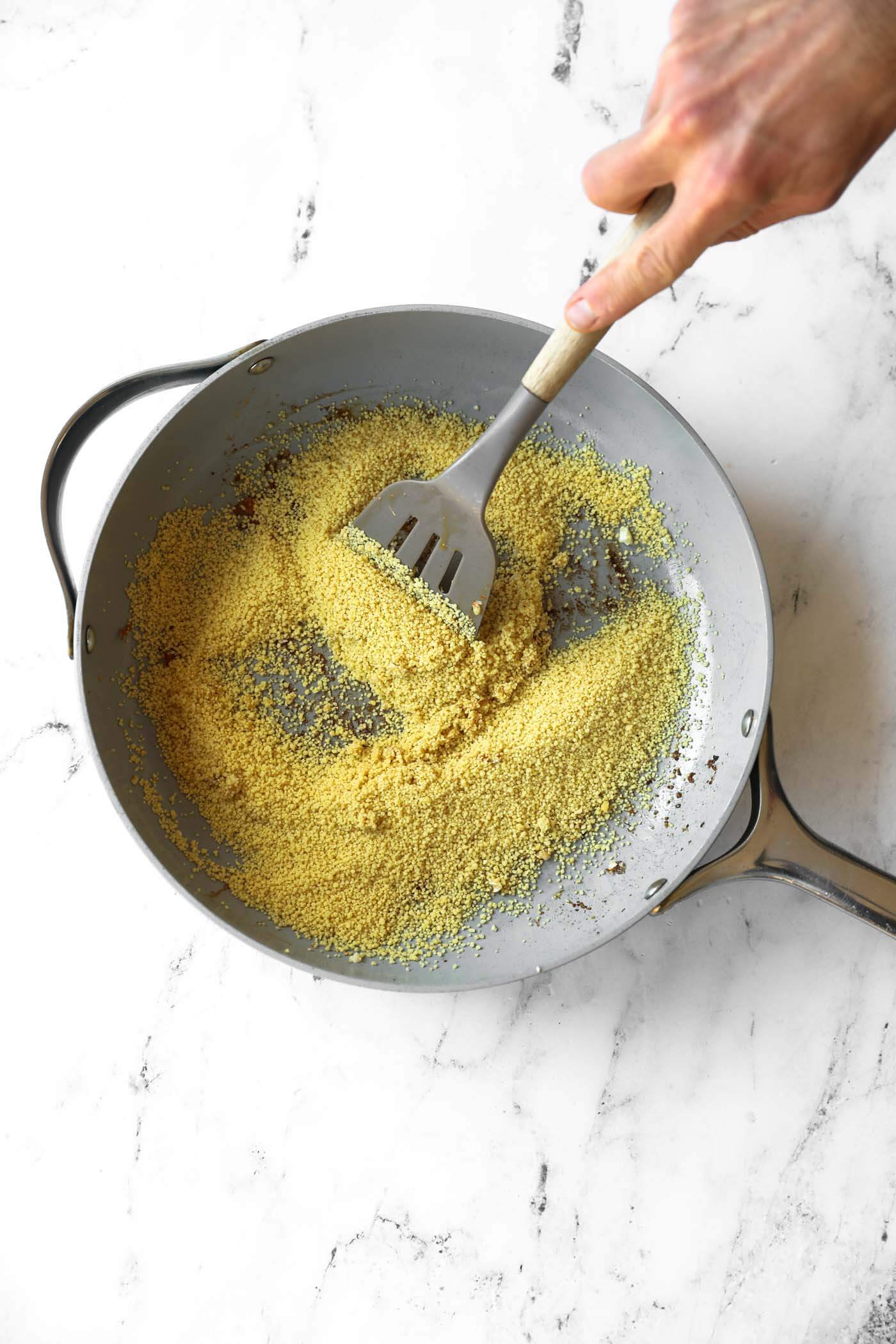 A hand stirs couscous in a large grey skillet with a slotted spatula on a white marble surface.