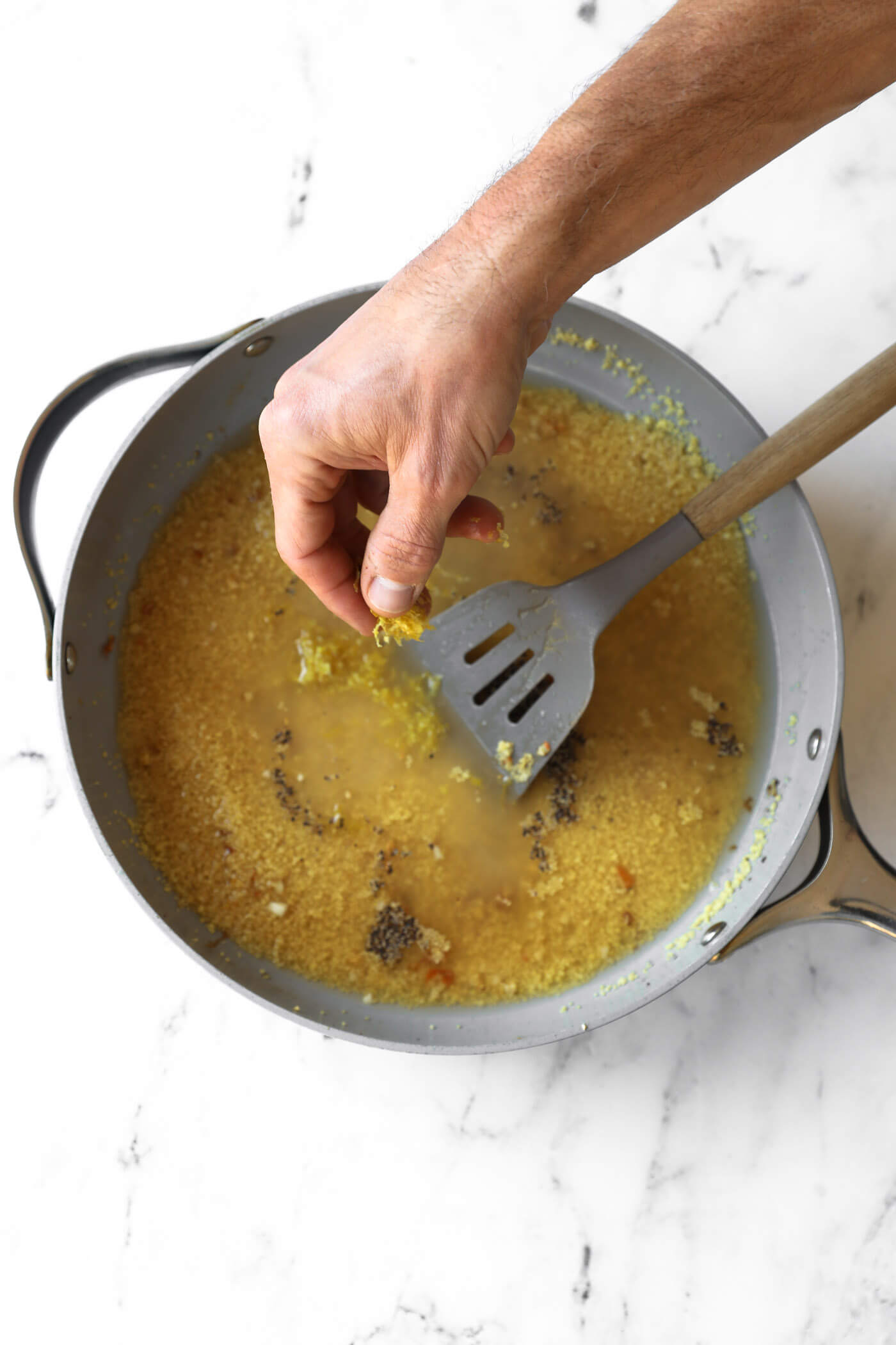 A hand sprinkles seasoning into a pan of couscous and broth, with a slotted spatula resting in the mixture on a white marble surface.