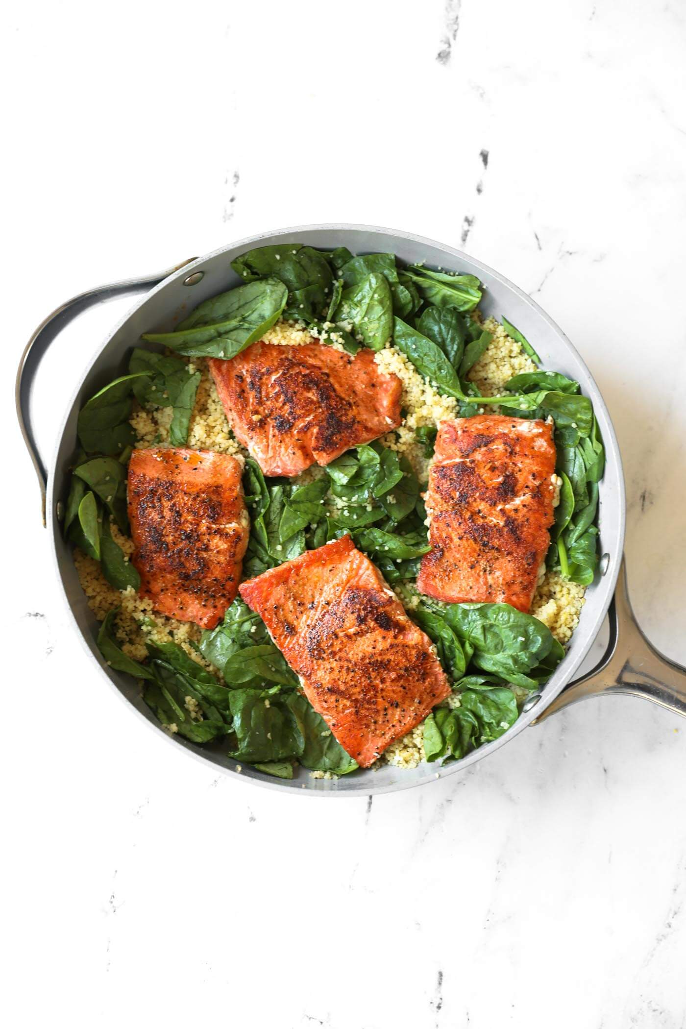 A skillet with four pieces of seasoned salmon on cooked quinoa and fresh spinach, set on a marble surface.