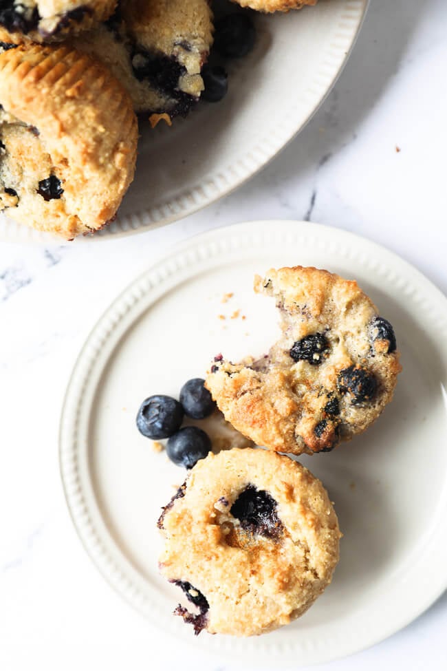 Vertical overhead image of a small plate with two gluten free blueberry muffins on it. One muffin has a bite taken out of it. 