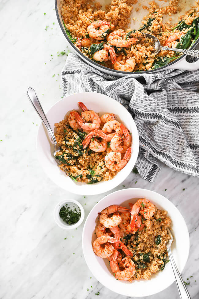 vertical overhead image of cajun shrimp and cauliflower rice in skillet and served up in two bowls with forks dug in. chopped parsley on the side. 