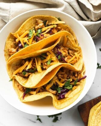 Overhead close up image of three tacos in a bowl. Ground chicken mixture, chopped cabbage, cilantro and shredded cheese on top.