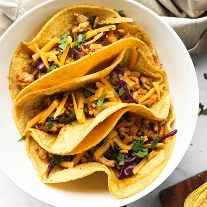 Overhead close up image of three tacos in a bowl. Ground chicken mixture, chopped cabbage, cilantro and shredded cheese on top.