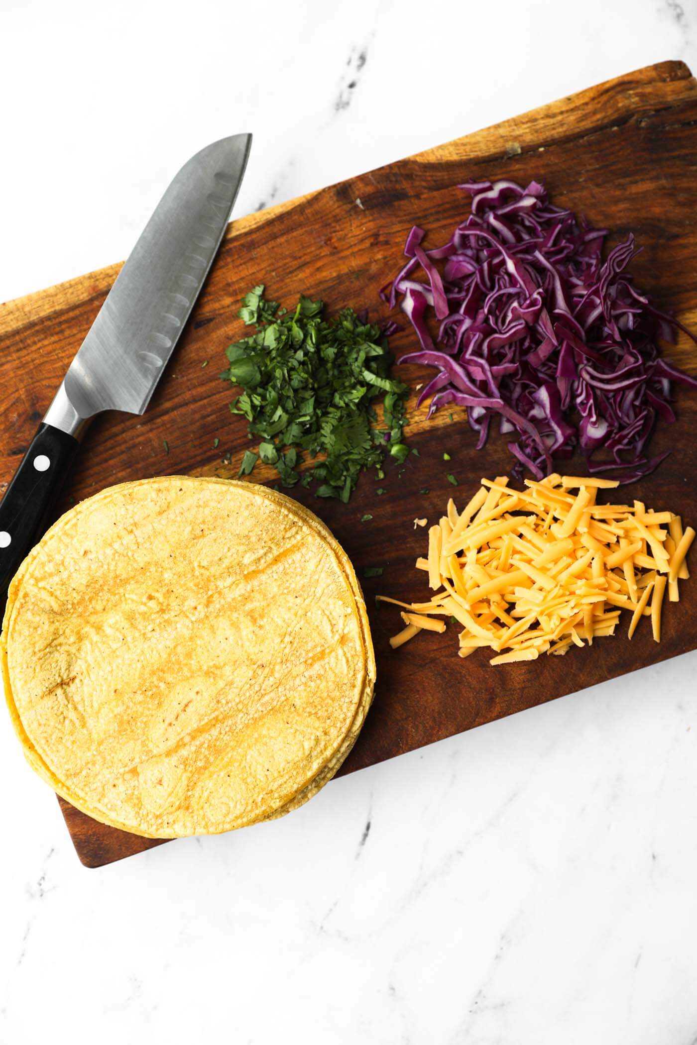 Overhead image of taco fixings on a cutting board - corn tortillas, grated cheese, chopped cabbage and cilantro.