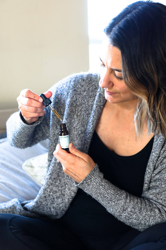 A woman sitting on a bed with a bottle of CBD oil with the dropper pulled out of the bottle. 