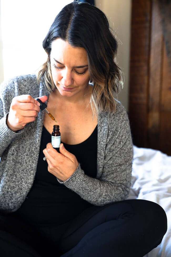 Woman sitting on a bed looking down as she pulls the dropper out of a bottle of CBD oil. 