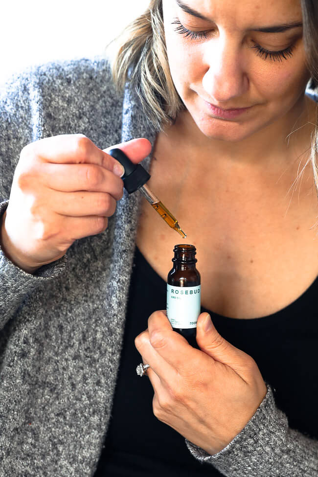 A close up of a woman sitting on a bed looking down at a bottle of CBD oil with the dropper pulled out. 