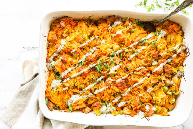 Overhead horizontal image of buffalo chicken casserole in casserole dish with chopped green onion and ranch drizzled on top and a serving spoon dug into dish. 