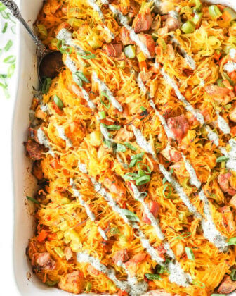 Overhead vertical image of buffalo chicken casserole in casserole dish with chopped green onion and ranch drizzled on top and a serving spoon dug into dish. Serving spoon dug into dish.