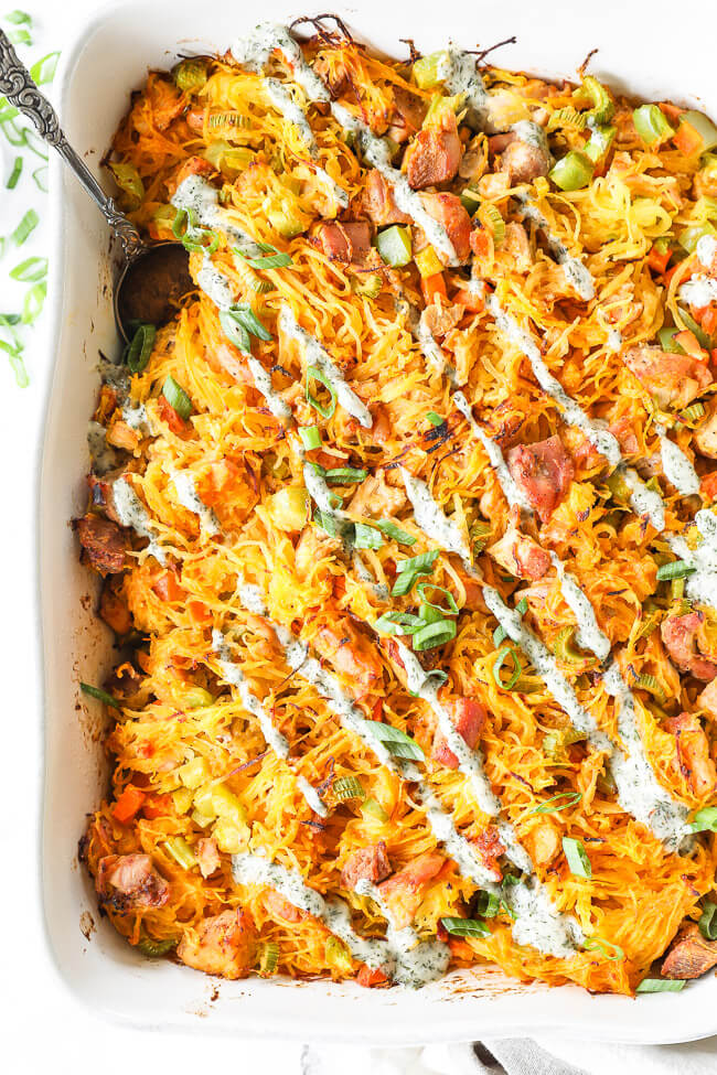 Overhead vertical image of buffalo chicken casserole in casserole dish with chopped green onion and ranch drizzled on top and a serving spoon dug into dish. Serving spoon dug into dish. 