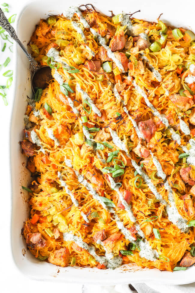 Overhead vertical image of buffalo chicken casserole in casserole dish with chopped green onion and ranch drizzled on top and a serving spoon dug into dish. Serving spoon dug into dish.