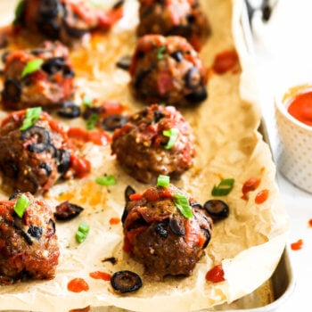 Angled image of pizza meatballs on a sheet pan with marinara sauce drizzled on top and garnished with chopped green onion.