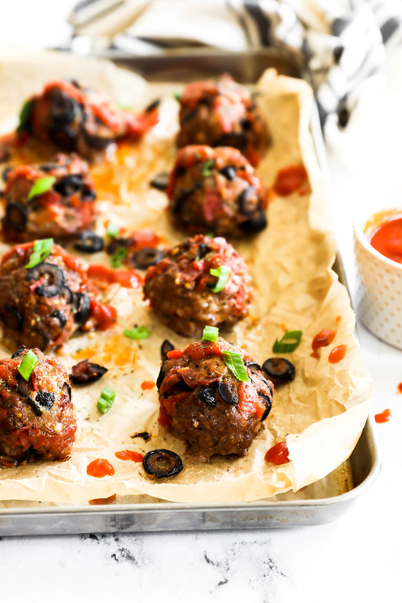 Angled image of pizza meatballs on a sheet pan with marinara sauce drizzled on top and garnished with chopped green onion.