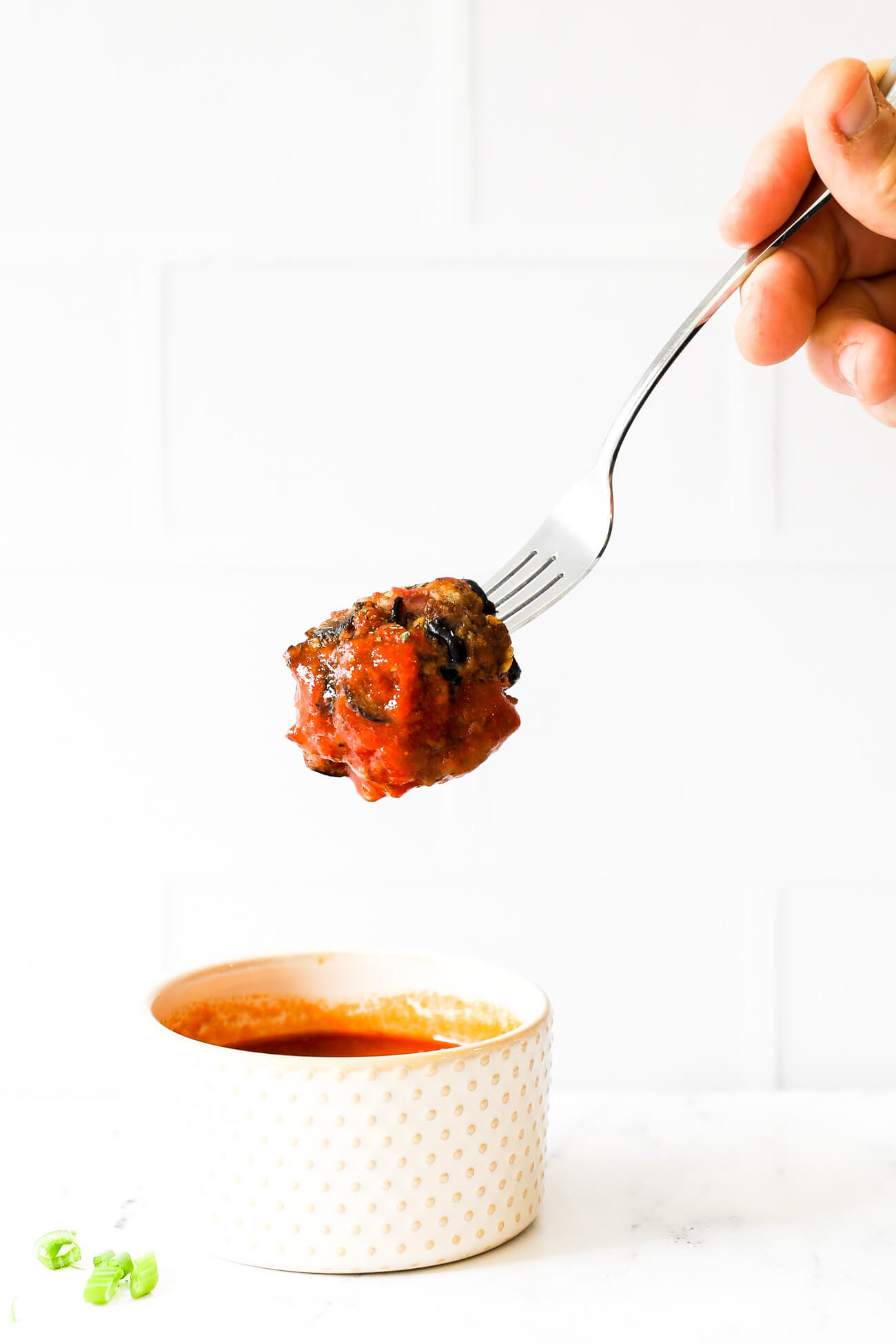 One pizza meatball on a fork being dipped into a ramekin of marinara sauce.