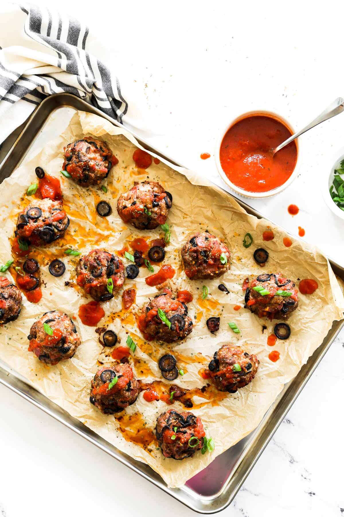 Overhead image of a sheet pan with pizza meatballs and marinara sauce on the sauce. Meatballs are drizzled with marinara sauce on top and sprinkled with chopped green onion.