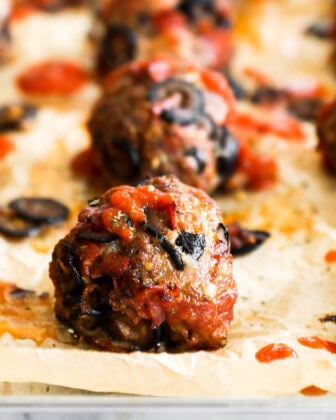 Close up of one pizza meatball on a sheet pan with more meatballs blurred out in the background. Meatball has marinara sauce drizzled on top.
