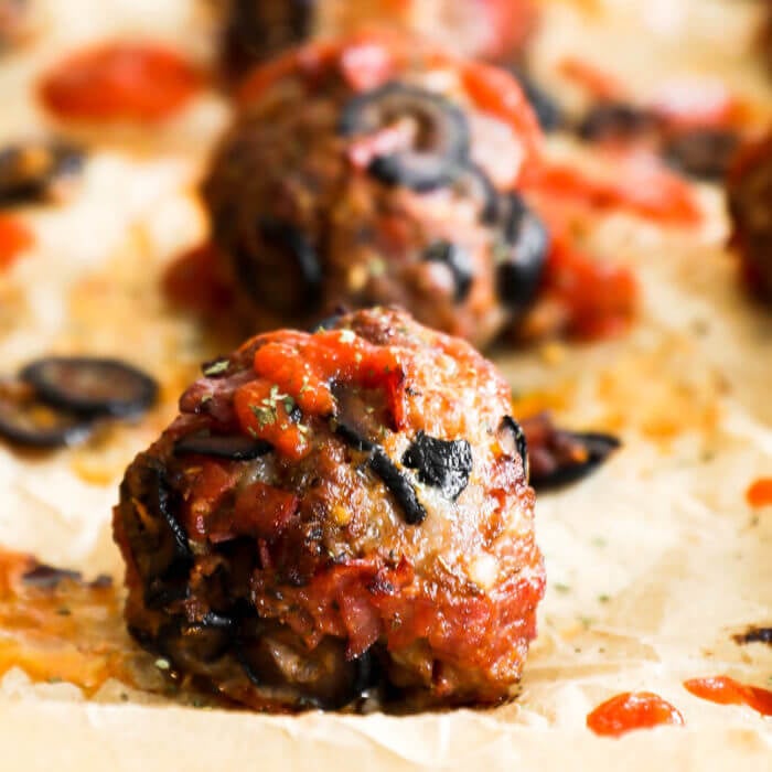 Close up of one pizza meatball on a sheet pan with more meatballs blurred out in the background. Meatball has marinara sauce drizzled on top.