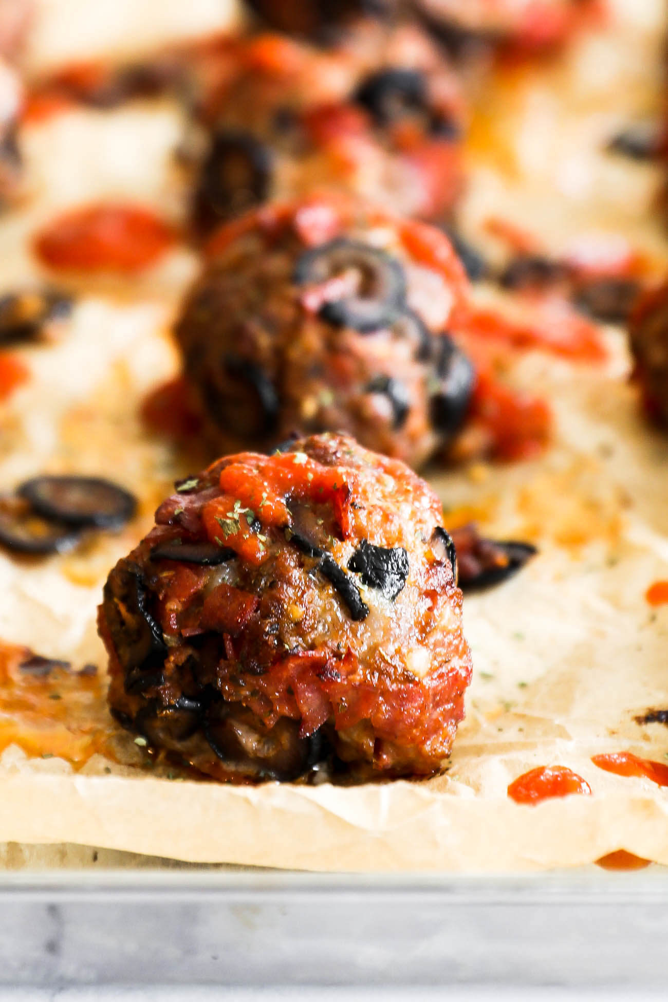 Close up of one pizza meatball on a sheet pan with more meatballs blurred out in the background. Meatball has marinara sauce drizzled on top. 