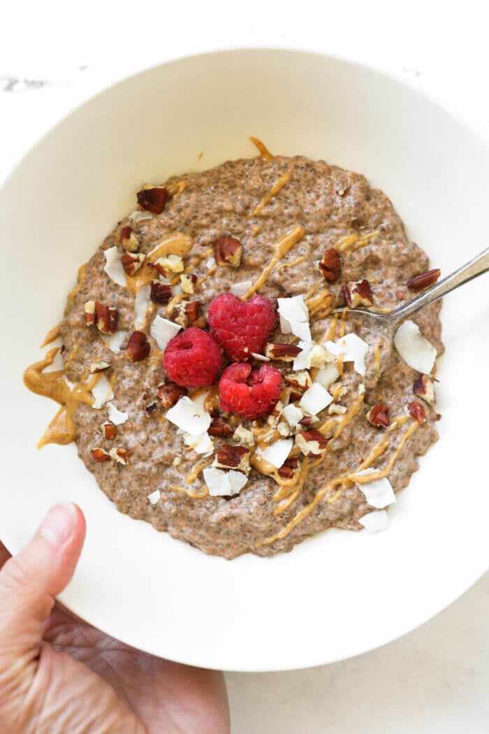 Close up image of holding a bowl chia pudding with a spoon in it. Toppings include shredded coconut, peanut butter, chopped pecans and a few raspberries.