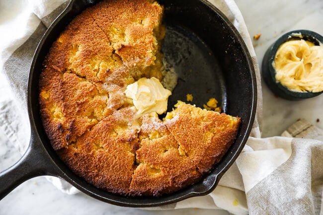 Paleo cornbread in a skillet with two pieces missing and whipped honey butter on top with more on the side. 