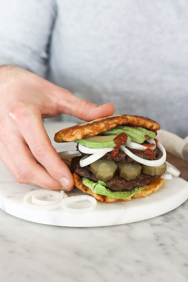 Paleo burger on board with avocado, onion, pickle and ketchup dripping down. Hand picking up burger vertical image.