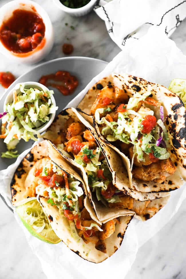Vertical overhead image of three fish tacos on a plate with extra coleslaw and salsa on the side. 
