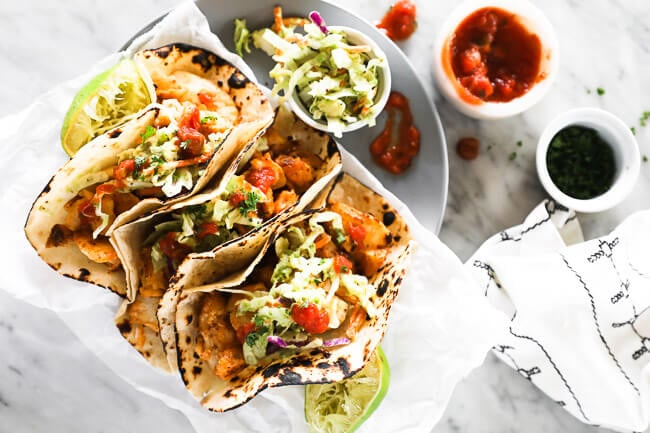 Horizontal image of three fish tacos on a plate with extra coleslaw and salsa on the side. 